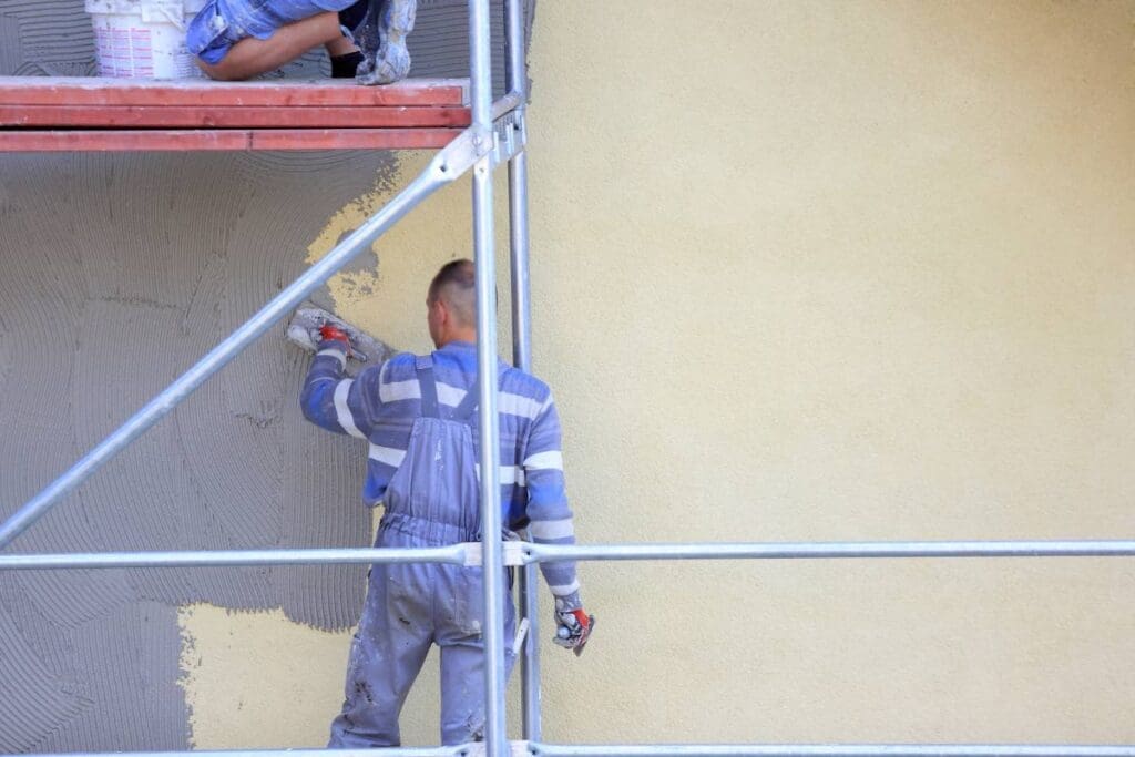 Pęknięcia tynku na elewacji domu – co może być przyczyną i jak naprawić? 2 Mężczyzna w kombinezonie remontuje elewację, nakładając nowy tynk